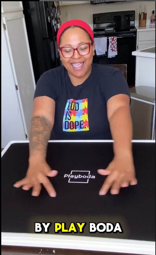 Happy user demonstrating the Playboda puzzle board in a bright kitchen, showing the black puzzle cover with Playboda logo.