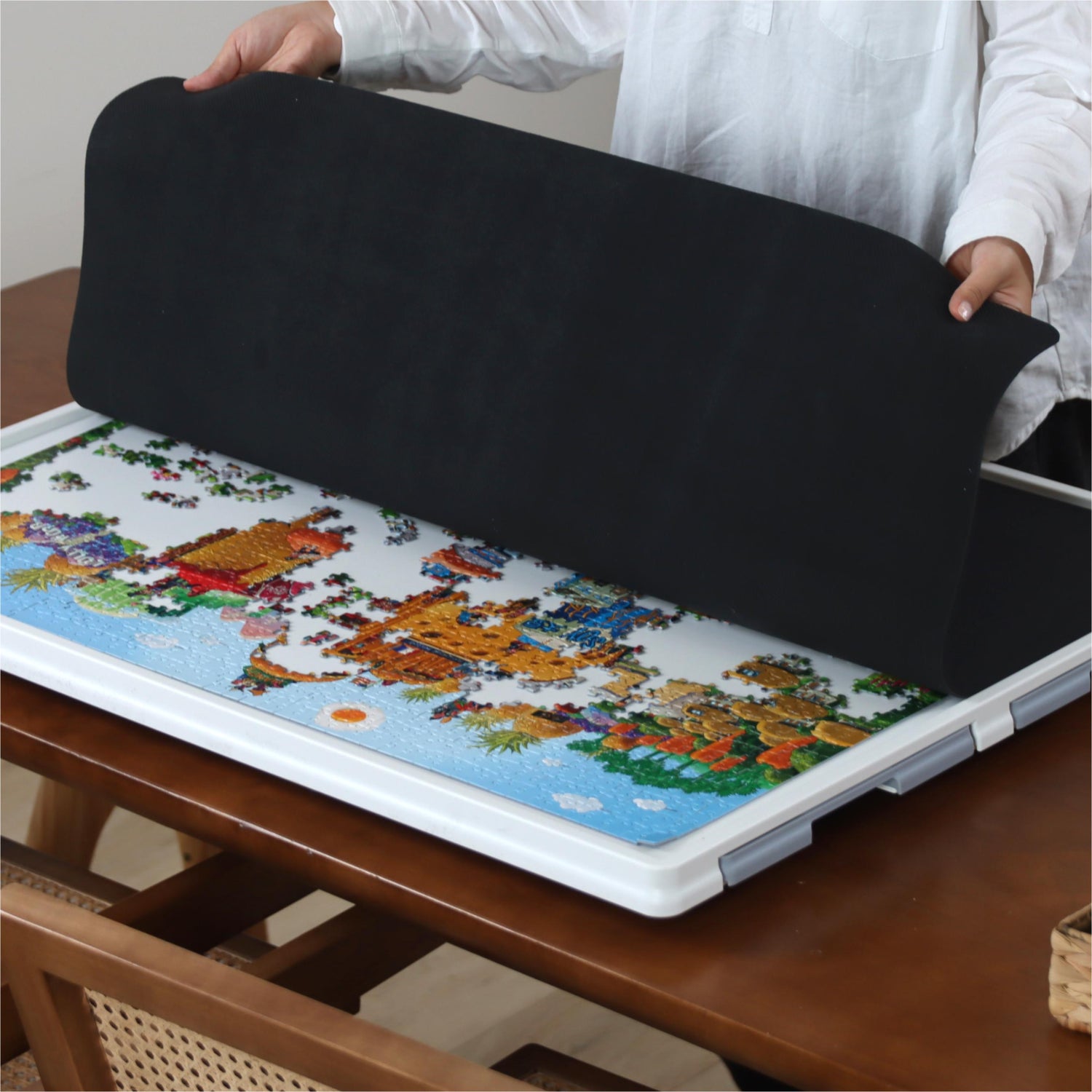Person covering a partially completed jigsaw puzzle with a black rubber mat on a white puzzle board to protect the pieces.