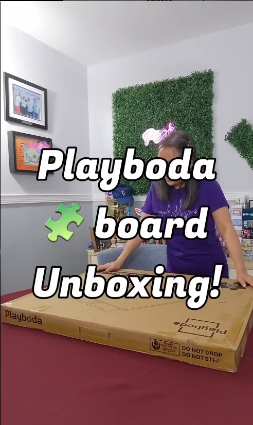 User unboxing a Playboda puzzle board package on a red table, showing the large shipping box with Playboda logo.